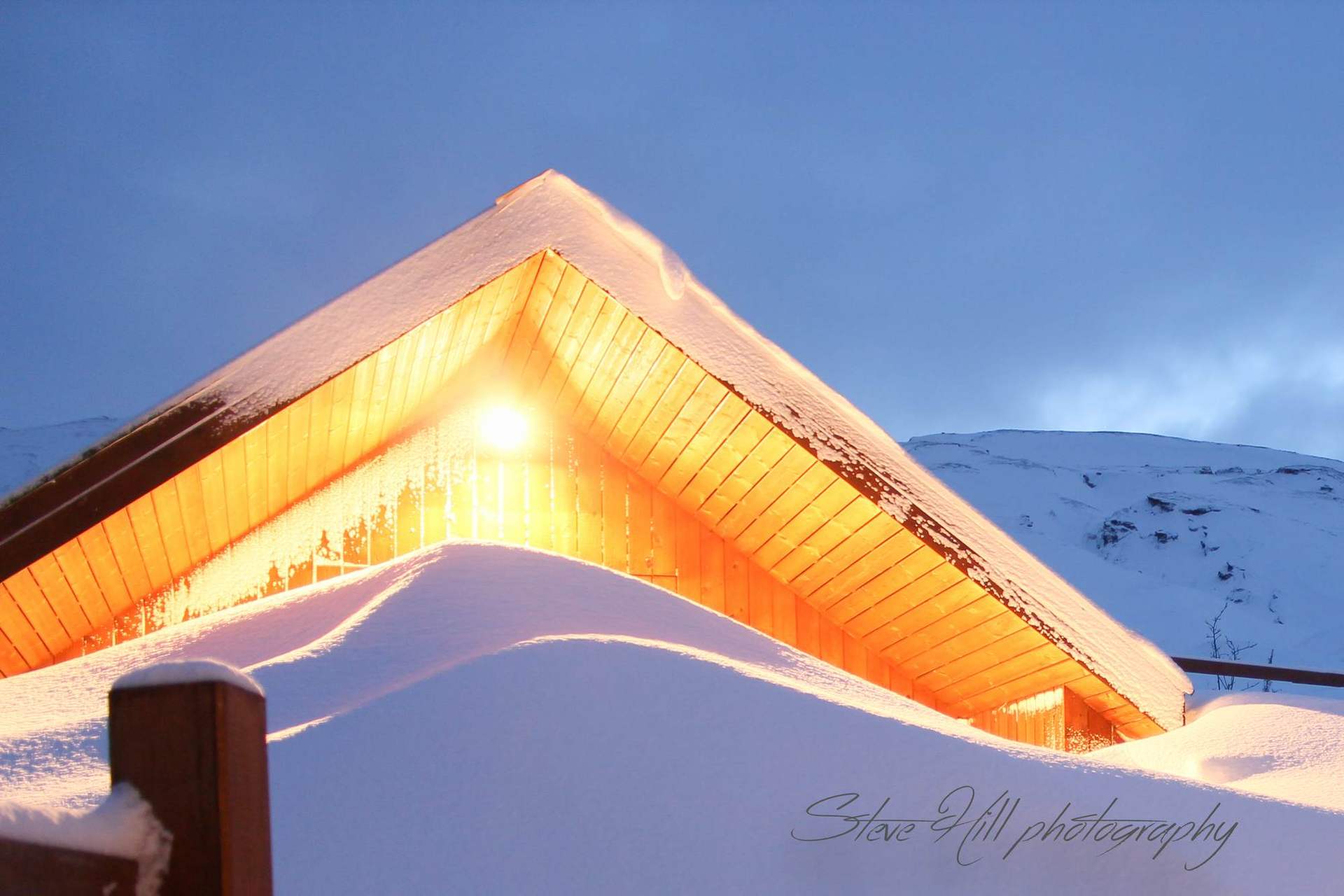 A log cabin half buried in snow at dusk, its roofline lit warm against a blue sky